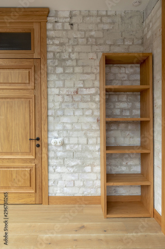 Closed oak panel door with black door knob beside empty bookcase against white brick wall with spots in light minimalist loft apartment