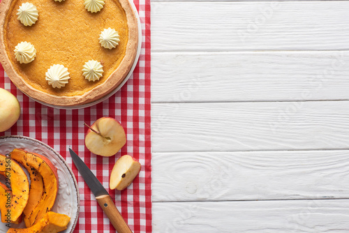 tasty pumpkin pie with whipped cream on checkered tablecloth near baked pumpkin, cut and whole apples, and knife on white wooden table