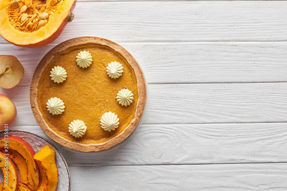 tasty pumpkin pie with whipped cream near baked and raw pumpkins, whole and cut apples on white wooden table