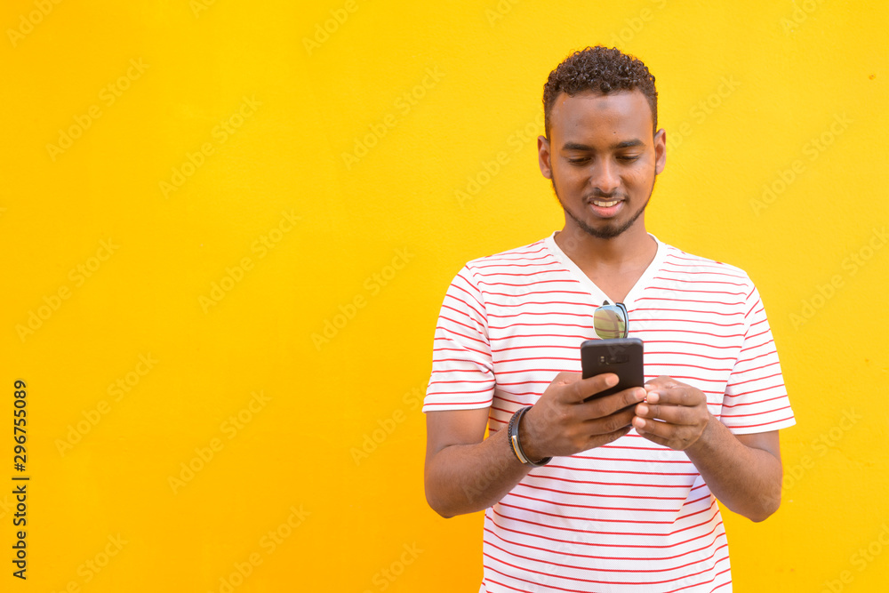Happy young African bearded man using phone against yellow wall Happy young African bearded man using phone against yellow wall