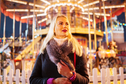Picture of young blonde woman in coat with fur collar in park on background of carousel
