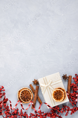 Christmas card with Holly berries, red ribbon, dried orange slices, cinnamon and white gift box on grey concrete background