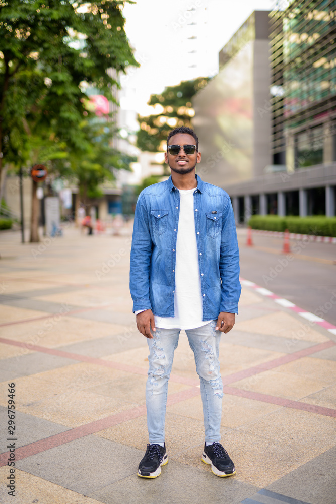 Full body shot of happy young African bearded man with sunglasses smiling in the city streets
