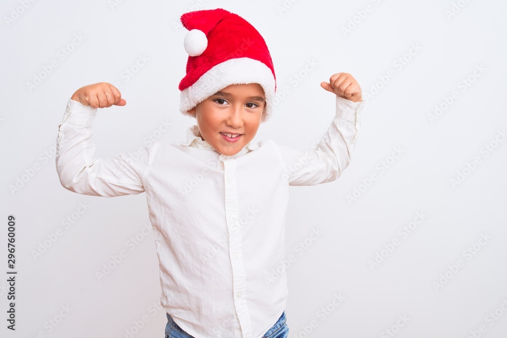 Beautiful kid boy wearing Christmas Santa hat standing over isolated white background showing arms muscles smiling proud. Fitness concept.