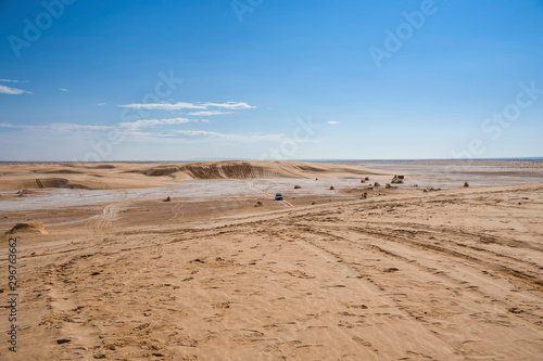 Sahara desert, with a salt marsh plot, with sand dunes, traces of past cars, and an off-road vehicle moving in the background