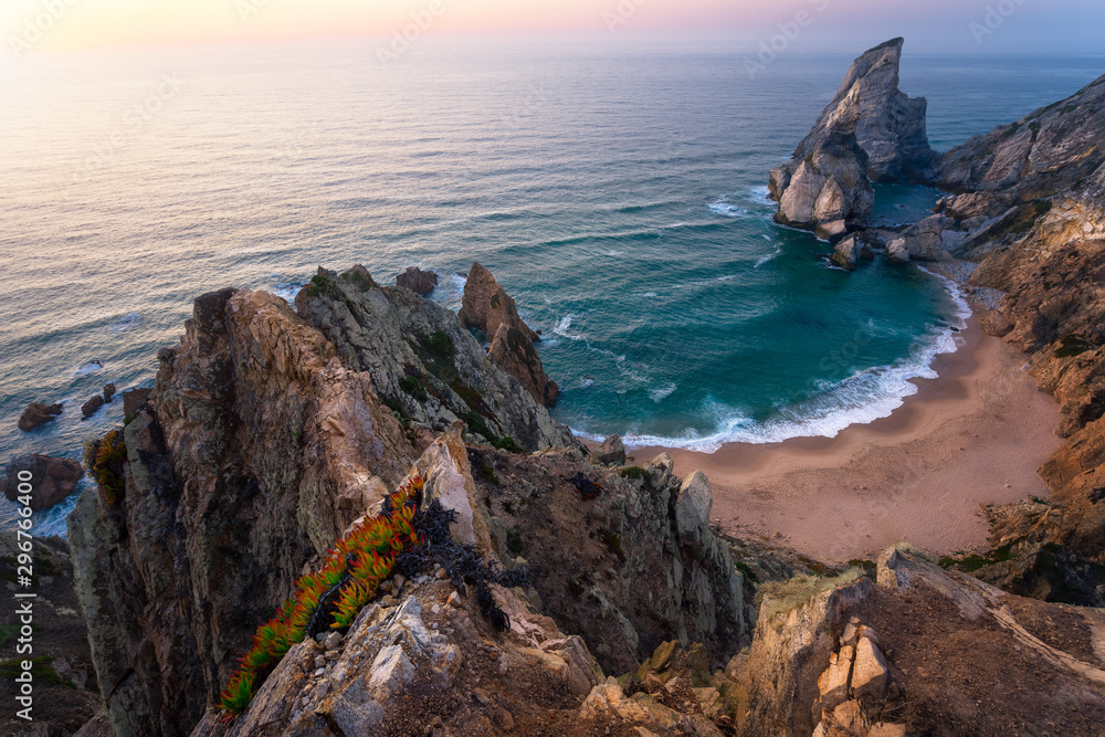 Praia da Ursa Beach from above. Rocky foreground with yellow flowers in sunset lit. Surreal scenery of Sintra, Portugal. Atlantic Ocean coastline landscape