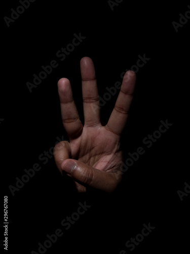 Low key photo of a brown man showing the number three with his fingers on a plain black background