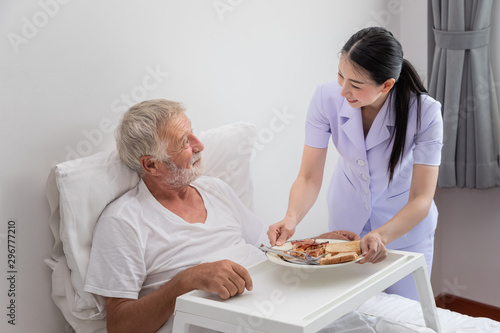 Happy nurse serving elderly senior man breakfast on bed in bedroom at nursing home