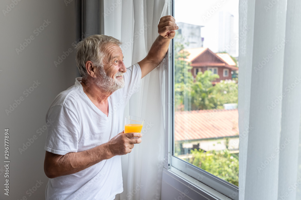 Senior elderly man standing and looking out of window in bedroom after waking up in morning with juice Senior elderly man standing and looking out of window in bedroom after waking up in morning with juice