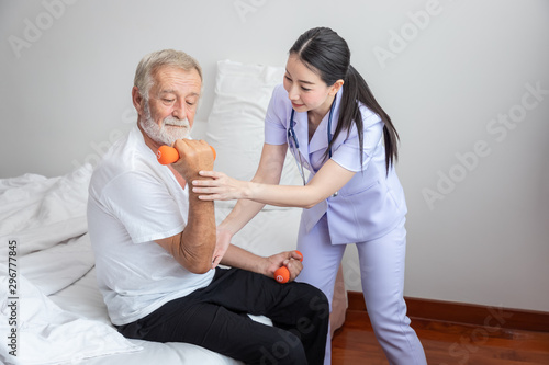 Senior man holding dumbbells with physiotherapist in bedroom sitting on bed, at nursing home