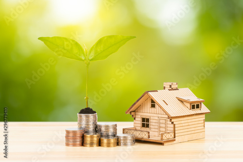 Coin stack and model house with growing leaves, on wooden desk on green tree background, mortgage concept