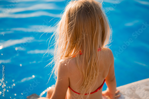 little blonde girl in a red swimsuit is sitting on the edge of the pool with her back to the camera