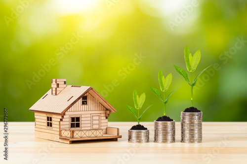 Coin stack and model house with growing leaves on wooden desk on green tree background, mortgage concept