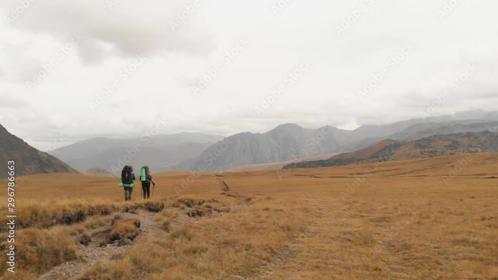 Aerial view of a couple of travelers man and woman with large backpacks in hats and sunglasses walk along an alpine plateau surrounded by epic mountains Aerial view of a couple of travelers man and woman with large backpacks in hats and sunglasses walk along an alpine plateau surrounded by epic mountains
