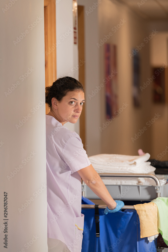 A serious and beautiful woman dressed in a white uniform, staring at the camera as she pushes a cleaning trolley in the hallway of a clinic. Healthy cleaning concept. A serious and beautiful woman dressed in a white uniform, staring at the camera as she pushes a cleaning trolley in the hallway of a clinic. Healthy cleaning concept.