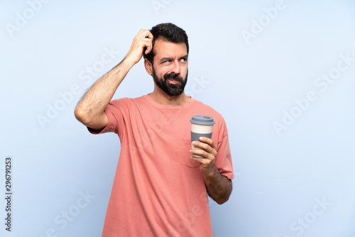 Young man with beard holding a take away coffee over isolated blue background having doubts while scratching head