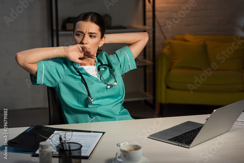 attractive nurse in uniform sitting at table and yawning during night shift