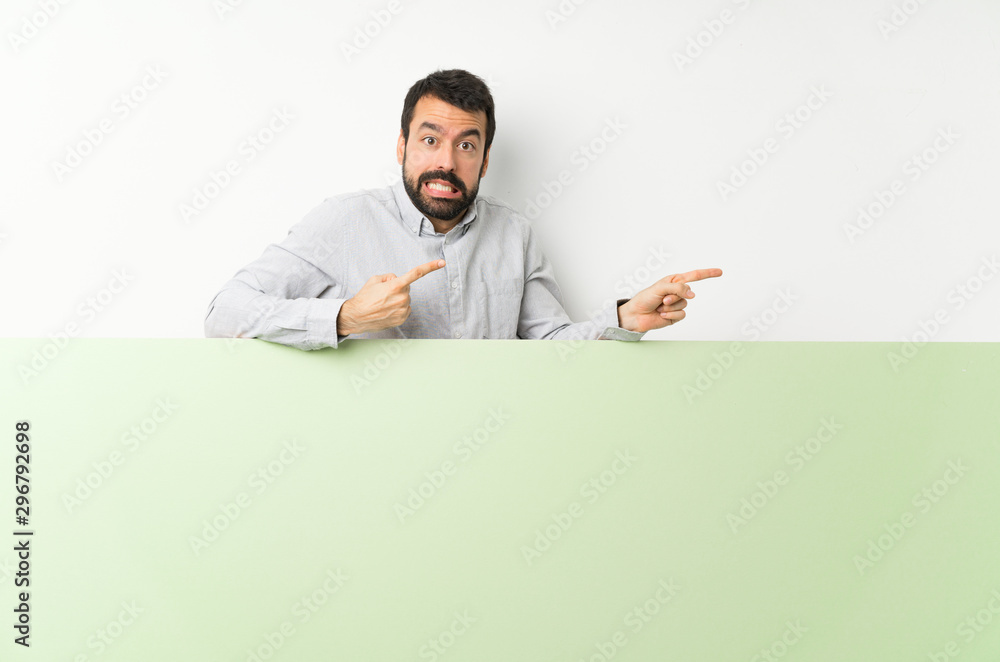 Young handsome man with beard holding a big green empty placard frightened and pointing to the side Young handsome man with beard holding a big green empty placard frightened and pointing to the side