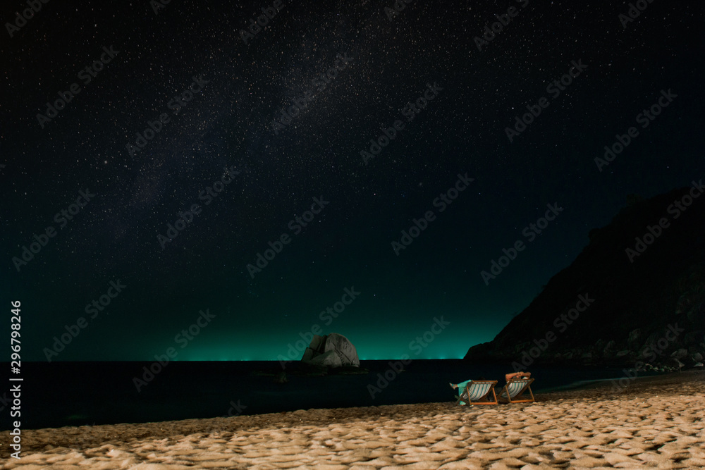 A couple admires the night starry sky in sun loungers on the beach. A quiet tropical night on the Paradise island of Koh Tao. The outline of the rocks above the water of the ocean. Starry sky over a