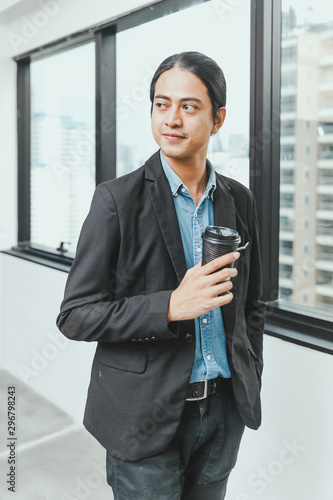 Portrait of handsome young businessman with long dark hair looking away while standing near the window