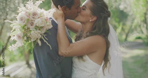 Happy multi ethnic couple kissing on their wedding day with a flower bouquet and green trees in the background with afternoon sun rays filtering through, the perfect wedding day