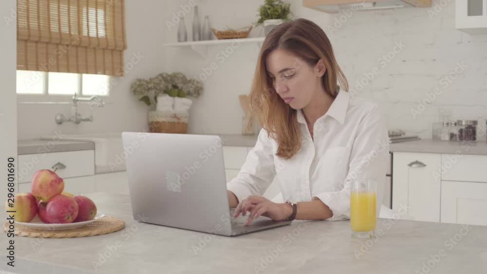 Young Caucasan businesswoman sitting at the table and typing on the laptop. Nice girl in white shirt working online from home. Young Caucasan businesswoman sitting at the table and typing on the laptop. Nice girl in white shirt working online from home.
