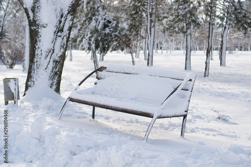 A view of the wooden snow-covered bench with a large layer of snow near snowy bushes and trees in winter park.