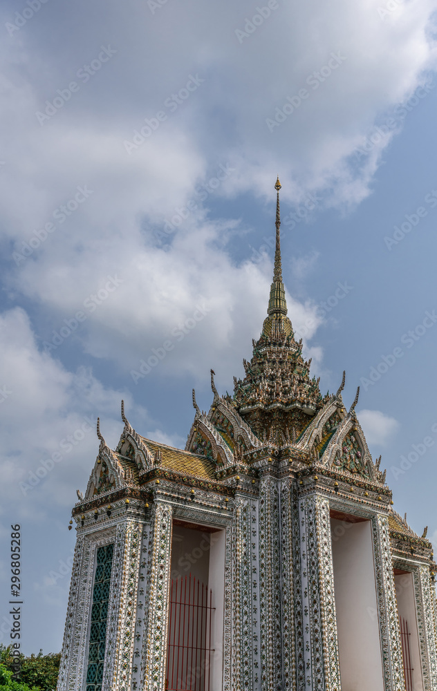 Fototapeta premium Bangkok city, Thailand - March 17, 2019: Facades, Gables and greenish spire of smaller section of Temple of Dawn, with its colorful porcelain faience surface, golden roof, against blue sky.