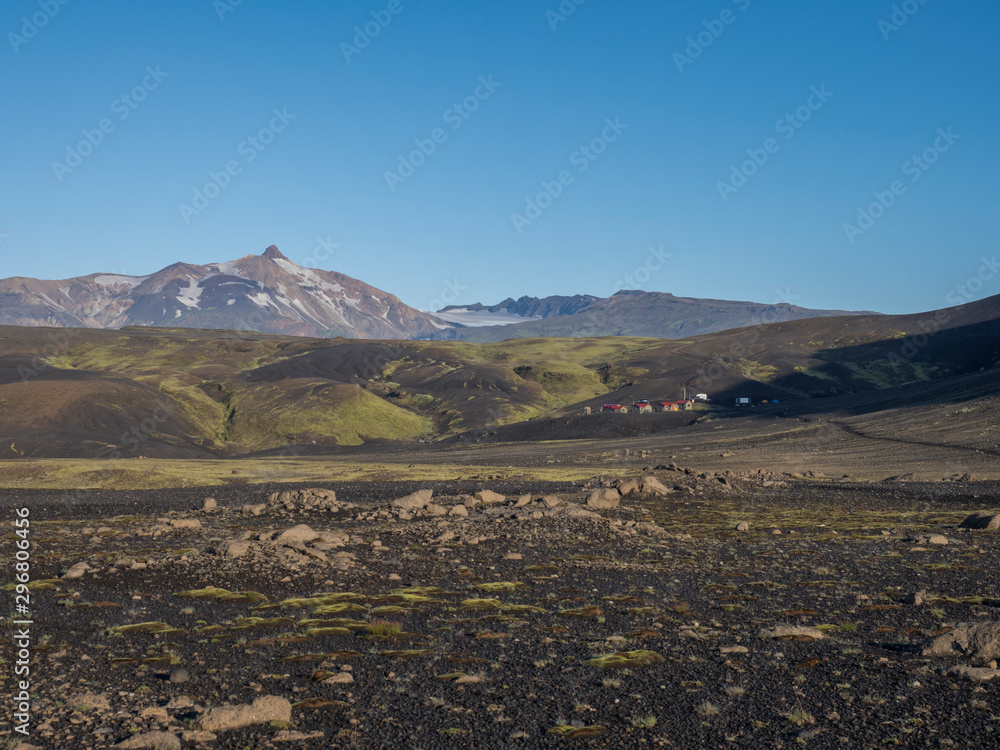 Fototapeta premium View on mountain hut at Iceland on Laugavegur hiking trail, green valley in volcanic landscape among lava fields. Early morning, summer blue sky