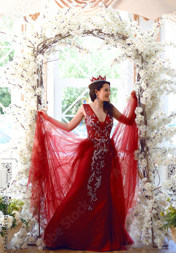 Portrait of happy young woman wearing red evening dress and tiara posing in arc with flowers. Pretty woman standing against window in studio