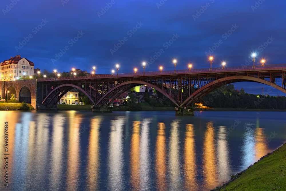 Wide angle landscape view of Old Bridge (also named the State) over Drava River. Light from lanterns reflected in water. Stunning autumn morning. Concept of landscape and nature. Maribor, Slovenia
