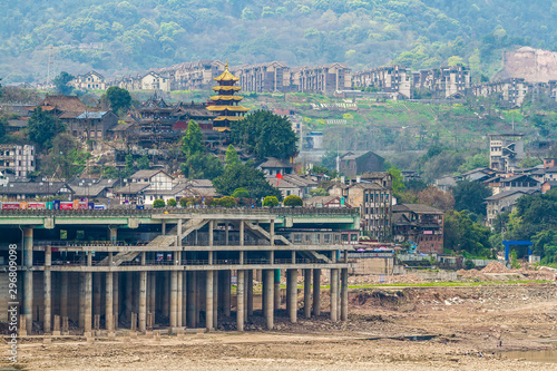 Chinese touristic quarter on the river bank.  Against the backdrop residential areas on the hills. Father and son fly a bright red kite on the dry bank of the Jialing River. Chongqing, China.