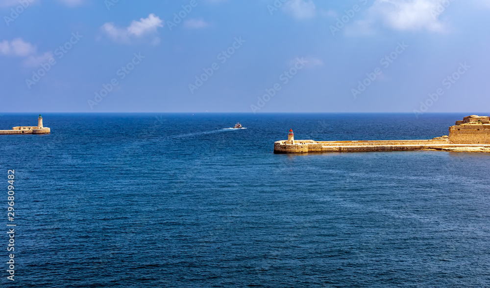 Boat going in turquoise sea water between Valletta breakwater with lighthouse and Ricasoli Breakwater Light at Grand Harbour East Breakwater at the entrance of the Grand Harbour.