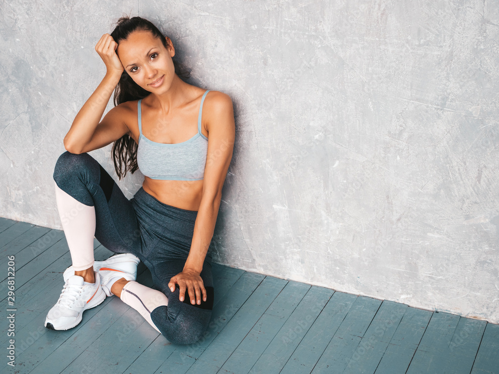 Fototapeta premium Portrait of confident fitness woman in sports clothing looking confident.Young female wearing sportswear. Beautiful model with perfect tanned body.Female sitting in studio near gray wall