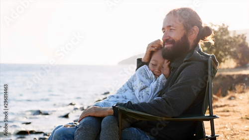 Cute child girl sits on hands of her father and they embracing. Happy family on vacation at lake shore, side view.