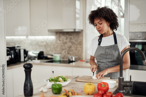 Gorgeous mixed race housewife in apron standing in kitchen and chopping mushrooms. On kitchen counter are all sorts of vegetables.