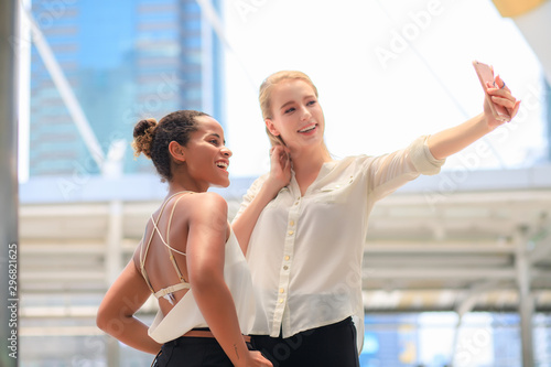 Two women wearing casual clothes are taking selfies. In the outdoor area in the city, there are tall buildings in the background during the day. © MPIX.TURE