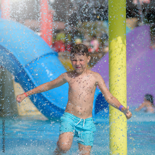 Joyful European boy in blue swimming shorts dancing in water park’s pool against colorful water slides, he smiling and enjoying his summer holidays. © Artem