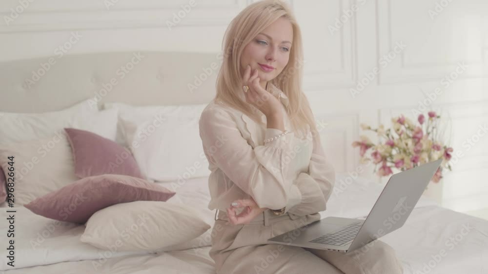 Smiling blond grey-eyed Caucasian businesswoman sitting with laptop on the bed and thinking. Young beautiful lady in official clothes working online from home. Smiling blond grey-eyed Caucasian businesswoman sitting with laptop on the bed and thinking. Young beautiful lady in official clothes working online from home.