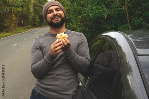 Young guy eating a burger near a car on an empty road. Food on the trip. Food on the go. Autumn travel. Fast food.