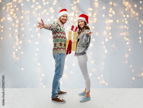 people and holidays concept - happy couple in santa hats with christmas gifts at ugly sweater party over festive lights background