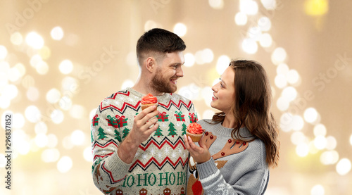 christmas, people and holidays concept - portrait of happy couple at ugly sweater party with cupcakes over festive lights background
