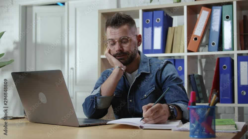 Bored hipster guy office worker suffering lack of motivation and interest in job profession sitting by table with laptop in office company. Bored hipster guy office worker suffering lack of motivation and interest in job profession sitting by table with laptop in office company.