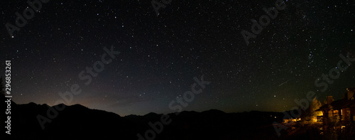 Shooting Star over cabin in the woods lit against starry night sky and shadowed mountain peak backdrop with distant light glowing beyond mountain panoramic landscape