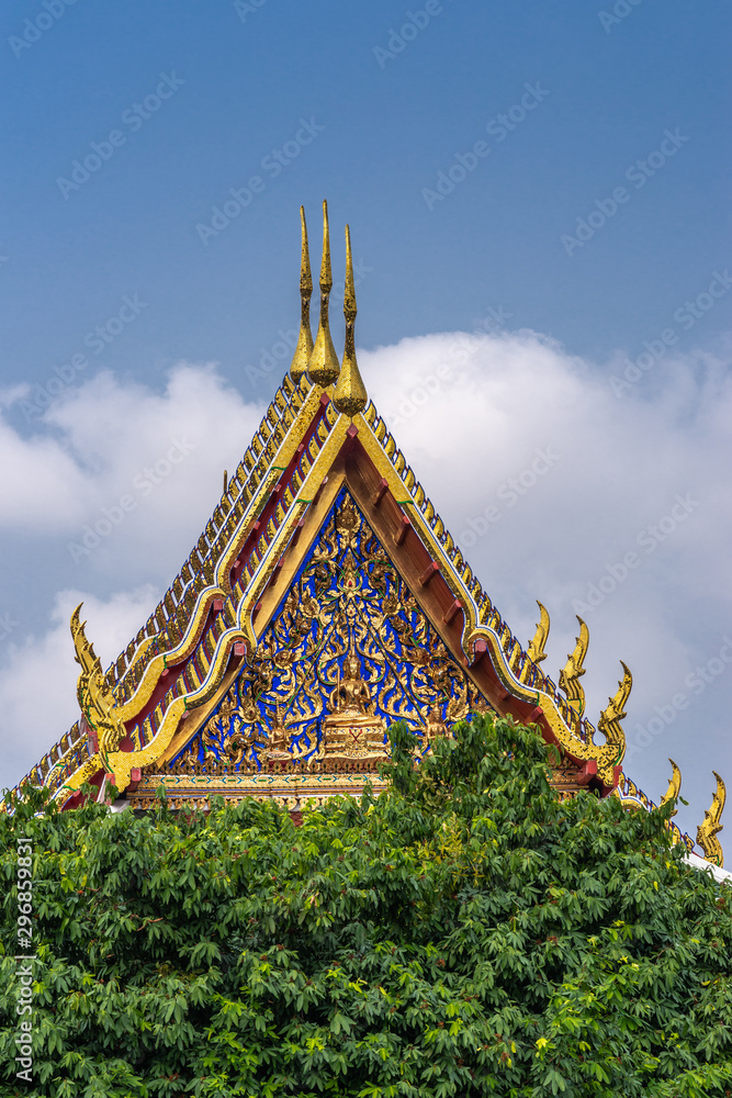 Fototapeta premium Bangkok city, Thailand - March 17, 2019: Three golden gables with intense blue decoration towering over green foliage at Temple of Dawn against blue sky.
