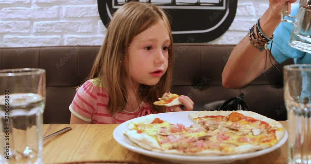 Portrait of six years old blonde girl eating pizza piece with hands, sitting in black couch next to her mother in a restaurant Portrait of six years old blonde girl eating pizza piece with hands, sitting in black couch next to her mother in a restaurant