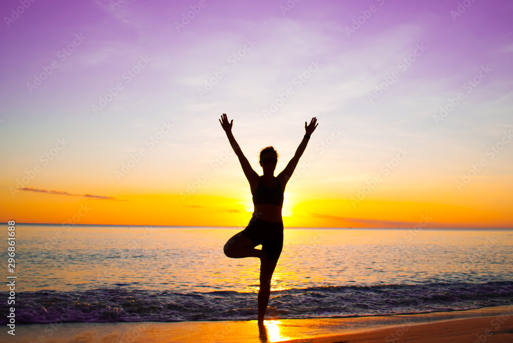 A young lady practices yoga on the beach in front of the setting sun. The Caribbean sea is tranquil as it laps the sandy coastline of the idyllic island A young lady practices yoga on the beach in front of the setting sun. The Caribbean sea is tranquil as it laps the sandy coastline of the idyllic island