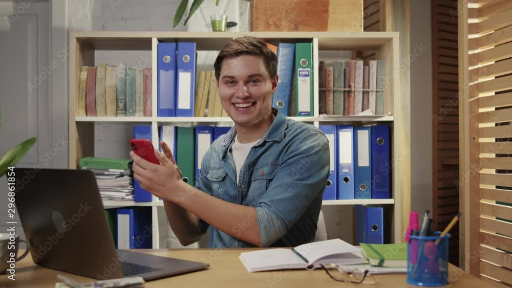 Joyful fun young business man employee having good time playing with smartphone typing on screen sitting at table in corporate office. Joyful fun young business man employee having good time playing with smartphone typing on screen sitting at table in corporate office.