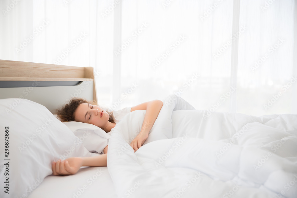 A beautiful white woman with curly hair is sleeping, resting and blanketing a bed in a white bedroom in her home in Thailand in the morning.