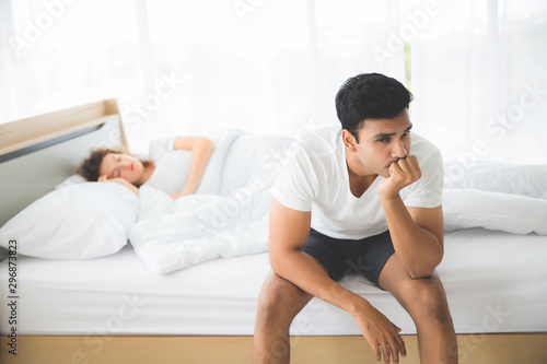 An Indian man was sitting. He had a strained facial expression and anxious. On the bed there was a sleeping woman.